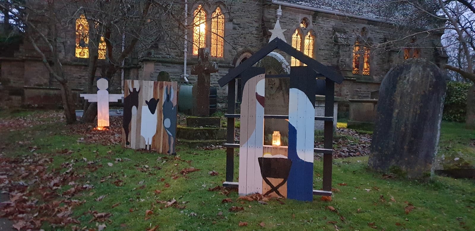 The handmade Nativity scene set out in St James' churchyard at dusk, the church windows glowing behind