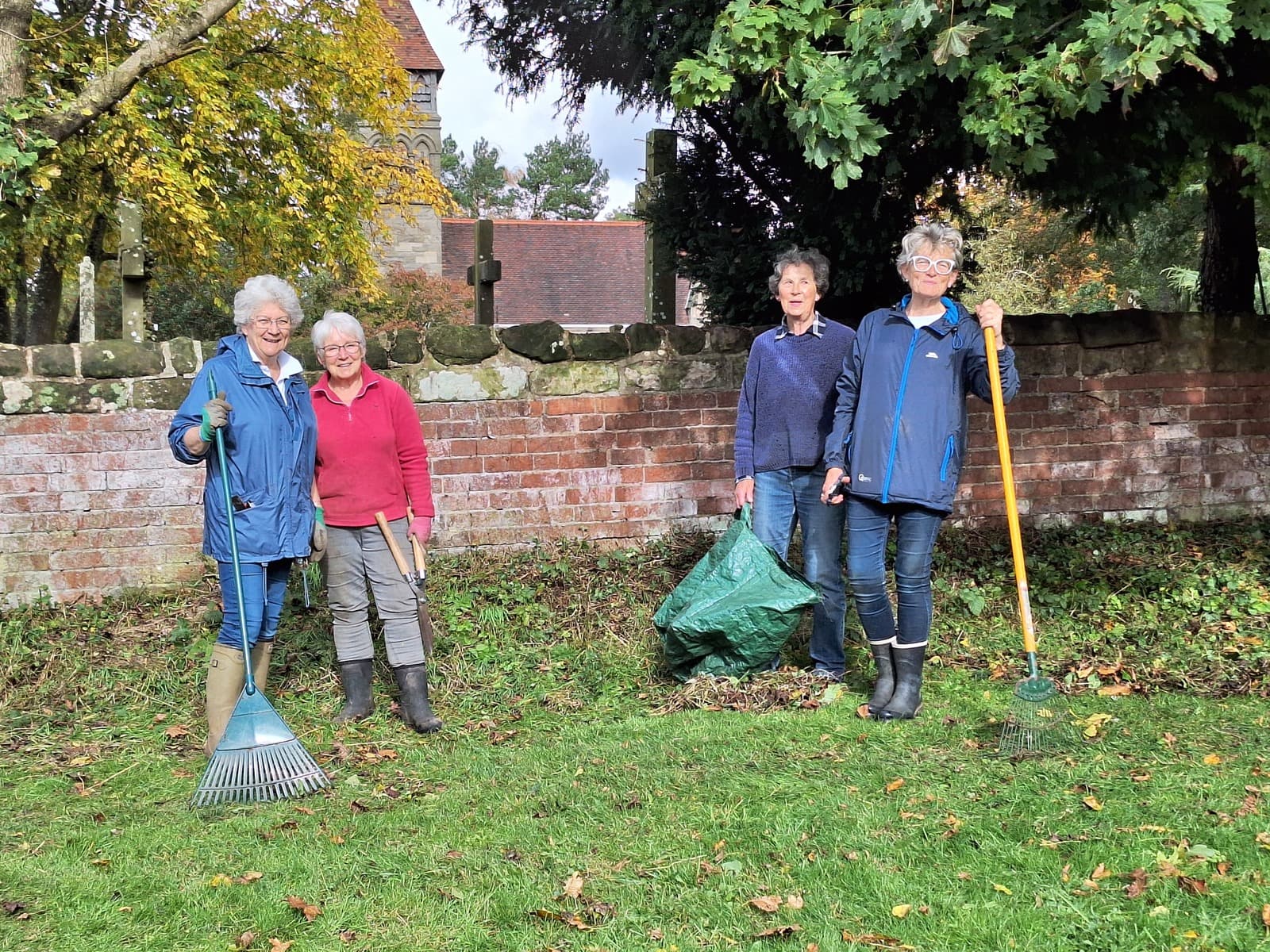 Four volunteers with rakes and a leaf bag during a churchyard clearing morning, the church and wall behind them